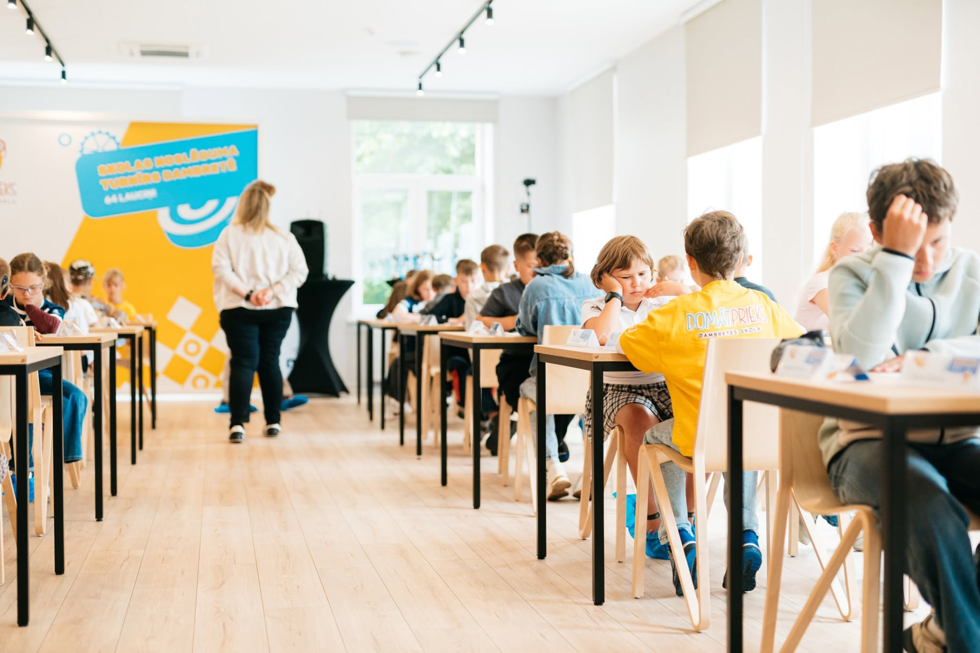 Dambretes turnīrs gaišā zālē ar dalībnieku galdiem